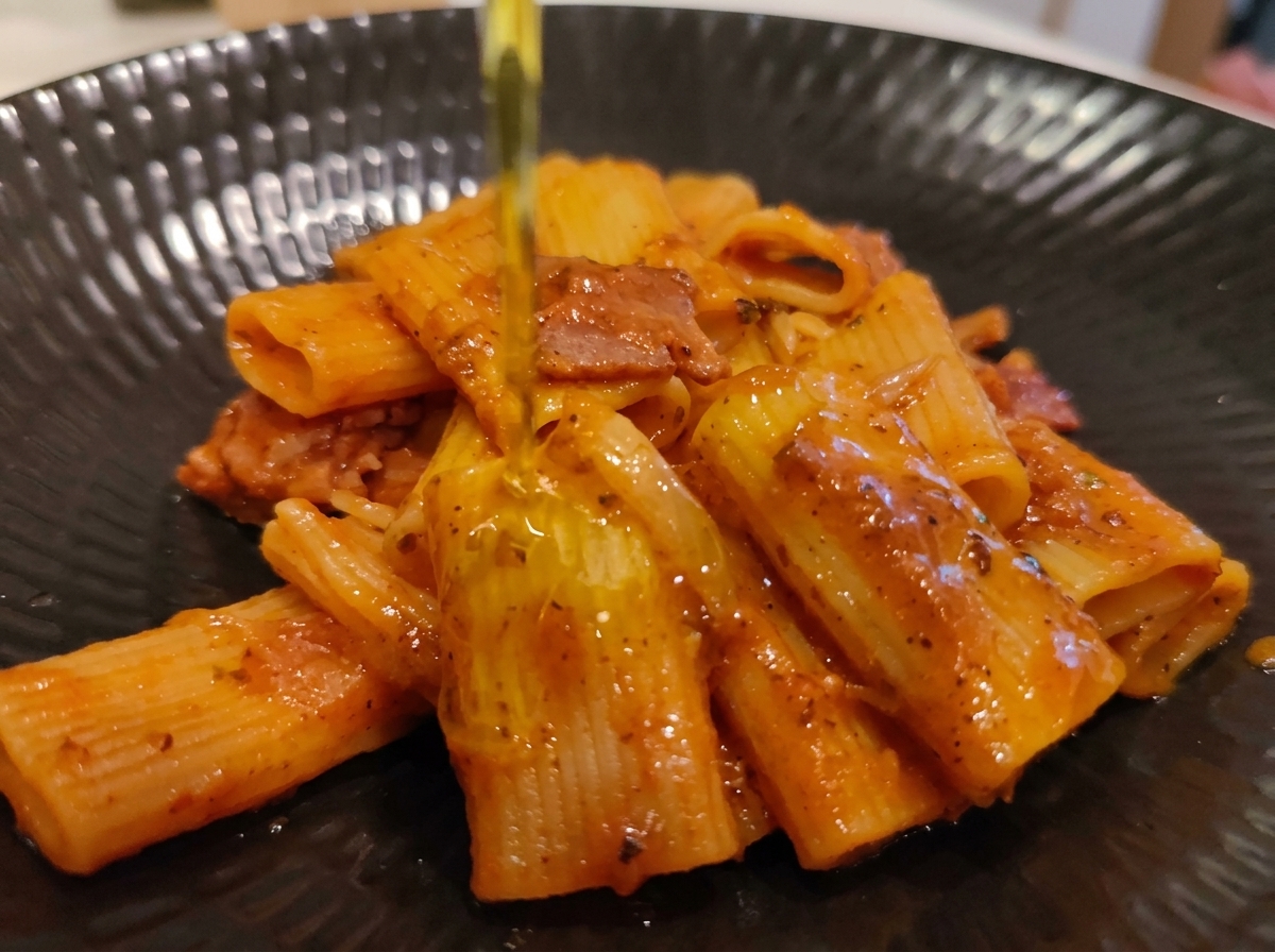 Pouring a stream of extra-virgin olive oil over a plated portion of rigatoni Amatriciana.