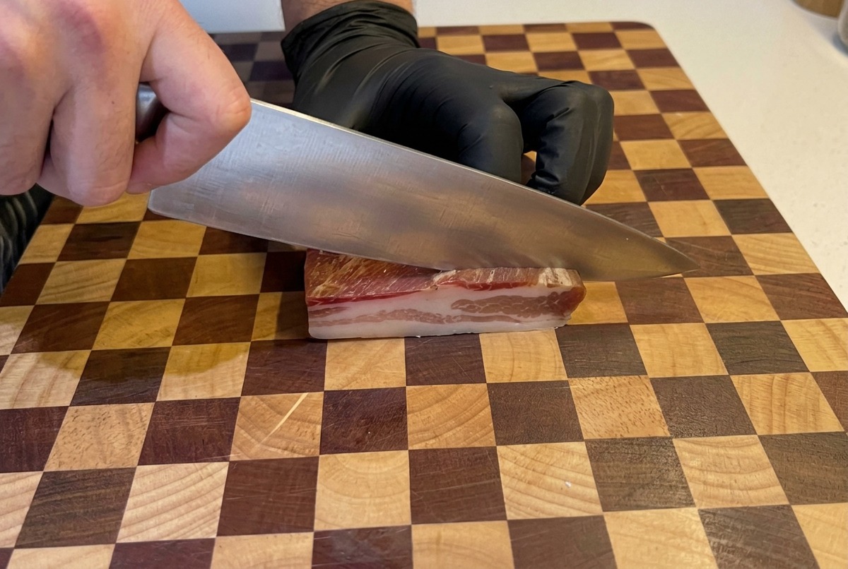 Hands wearing black gloves use a large chef knife to slice a thick slab of pancetta into small cubes on a checkered wooden board.