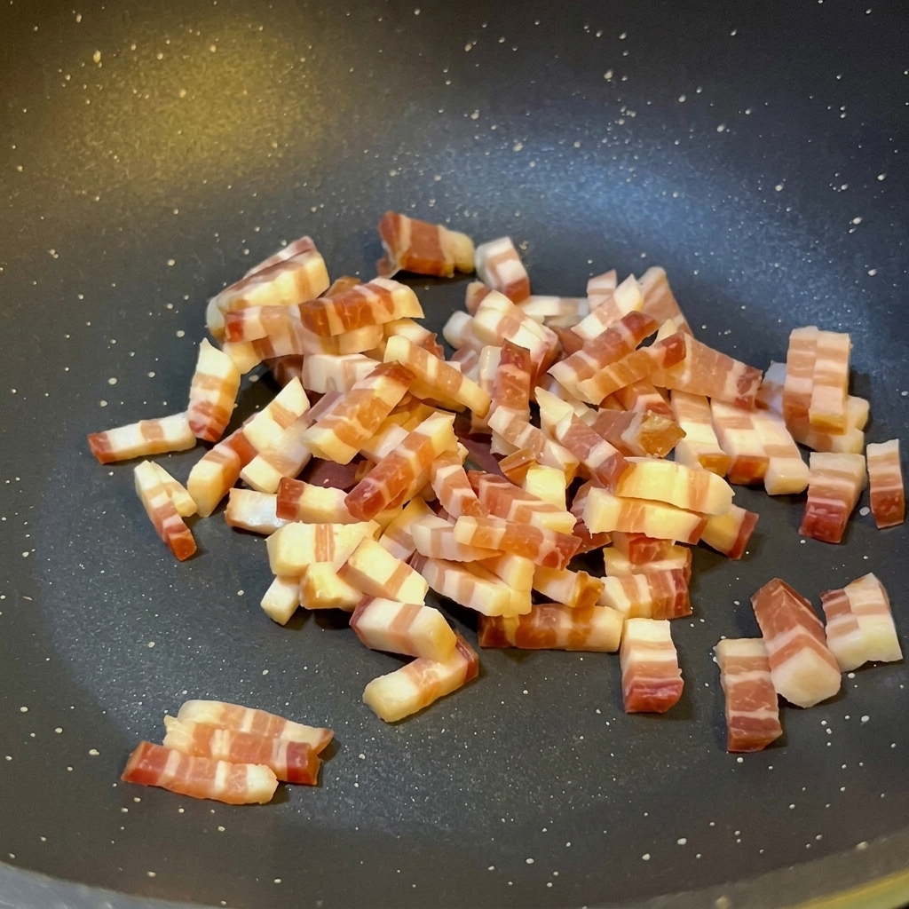 Small cubes of Italian pancetta being sautéed in a dark non-stick pan, showing some rendered oil and light browning on the edges.