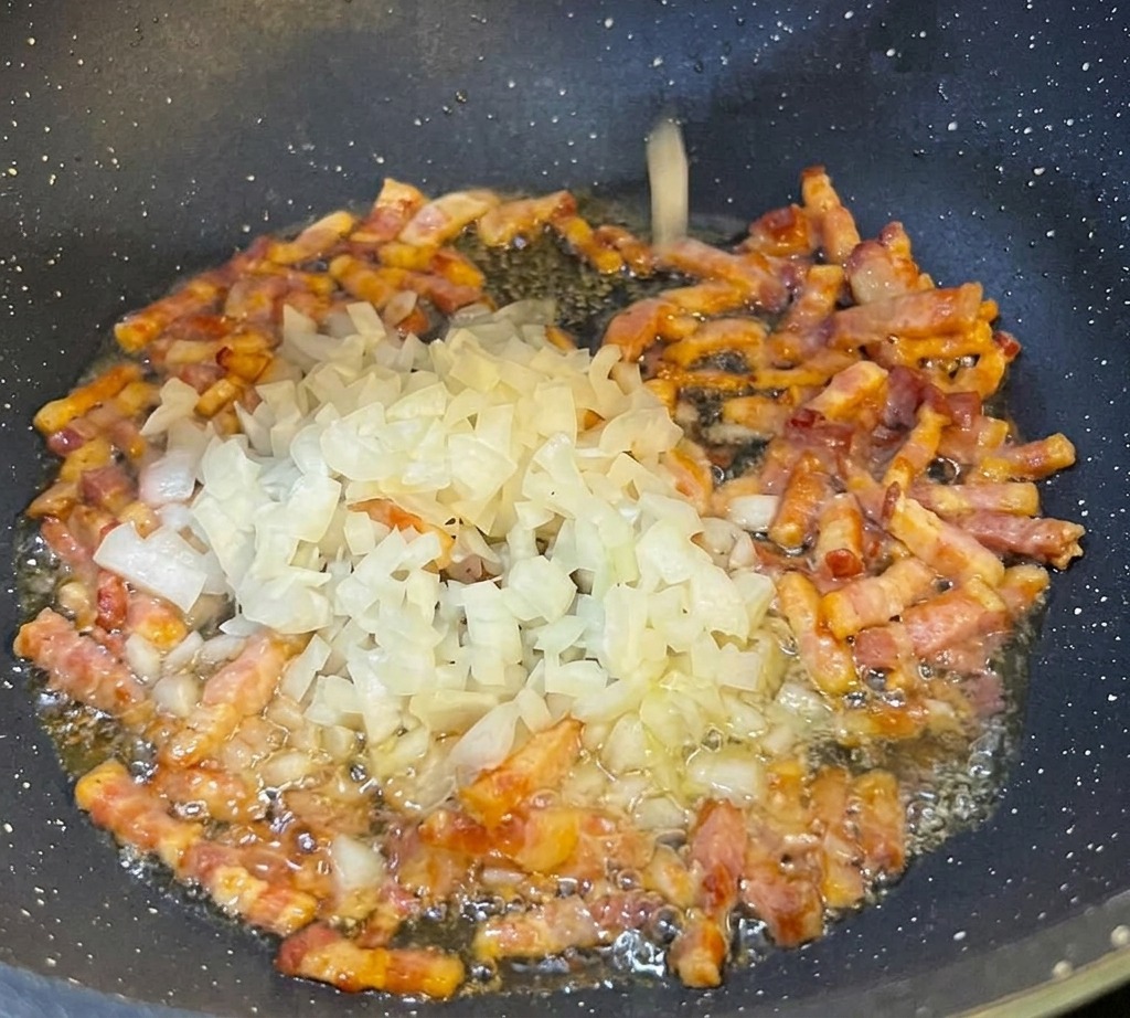 Finely diced white onions being sprinkled into a black frying pan containing browning strips of pancetta.