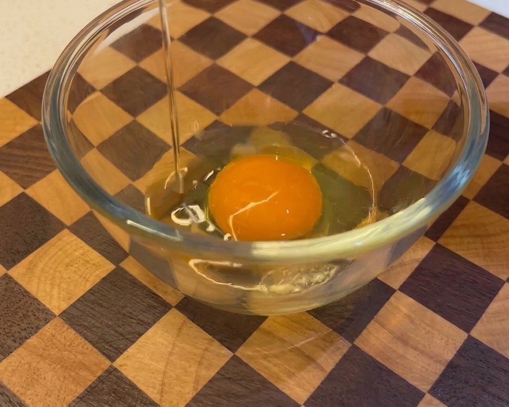 A raw egg being cracked and poured into a glass bowl that already contains one egg yolk, set against a wooden background.