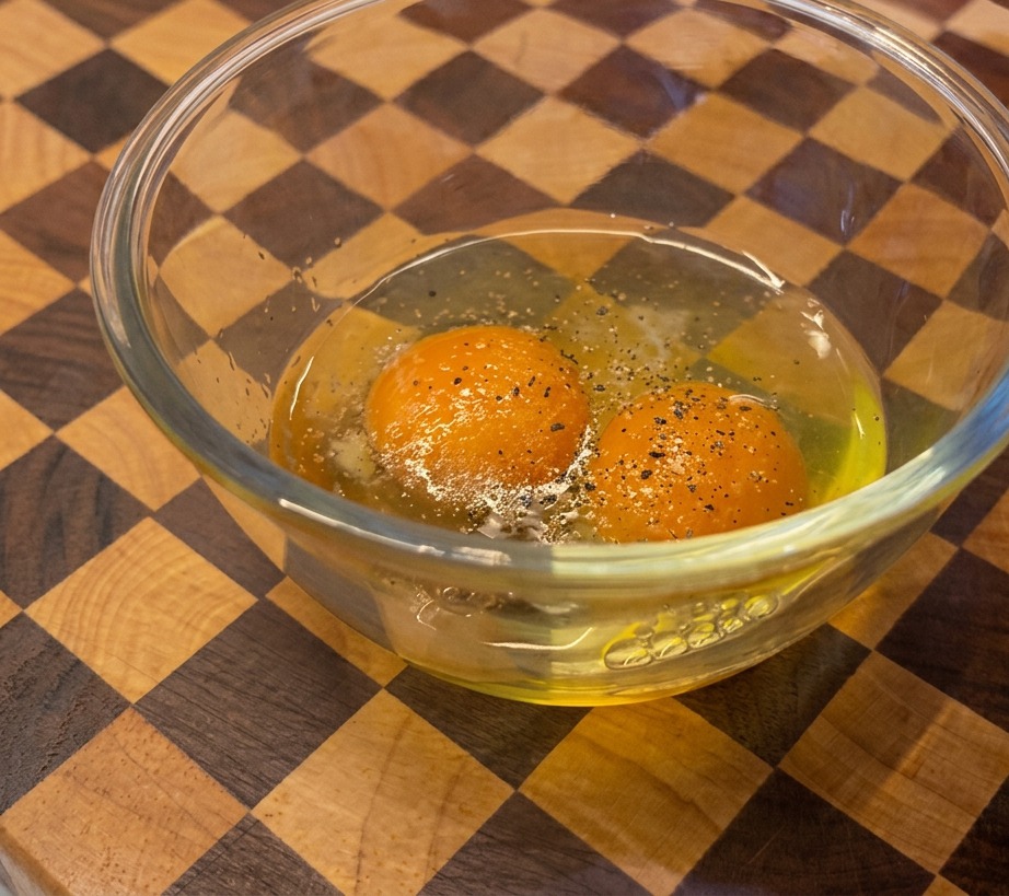 A person grinding black pepper from a wooden mill into a glass bowl containing two bright orange egg yolks.