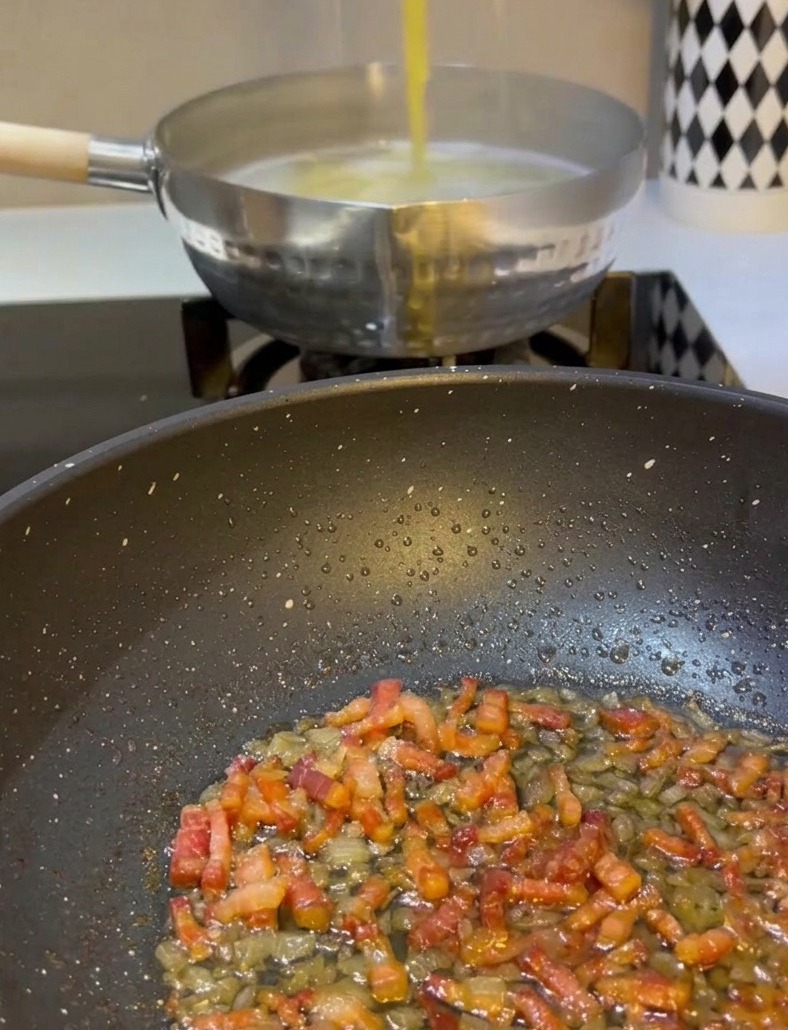 A hand using metal tongs to lift yellow spaghetti from a pot of boiling water and transferring it into a frying pan containing fried pancetta and onions.