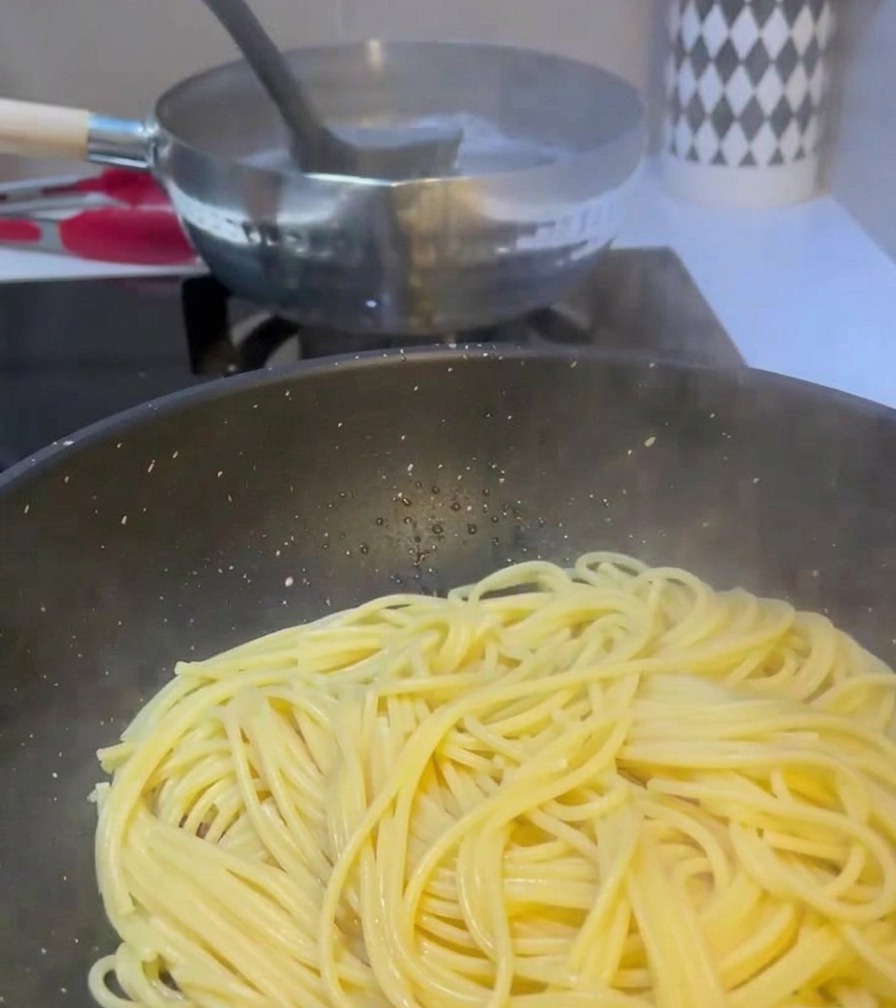 A ladle transferring cloudy water from a boiling pot of pasta into a pan already containing spaghetti and cooked meat.