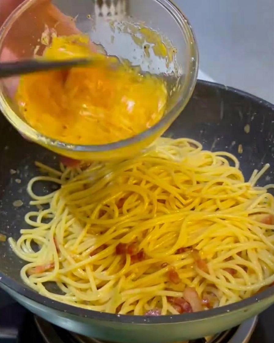 Pouring a vibrant yellow mixture of eggs and cheese from a glass bowl onto a pan of hot spaghetti and fried meat.