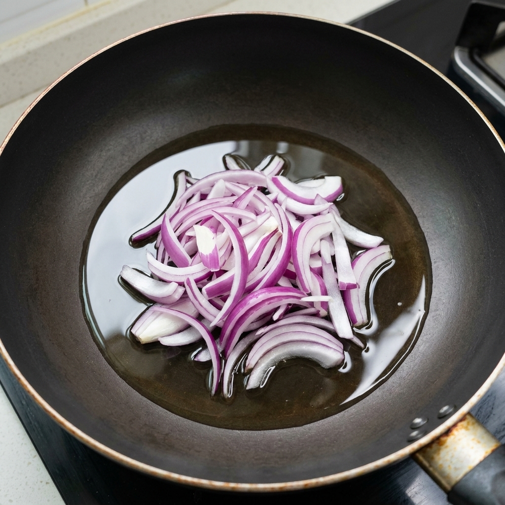 Sliced red onions being stir-fried in a shallow layer of oil in a black pan.