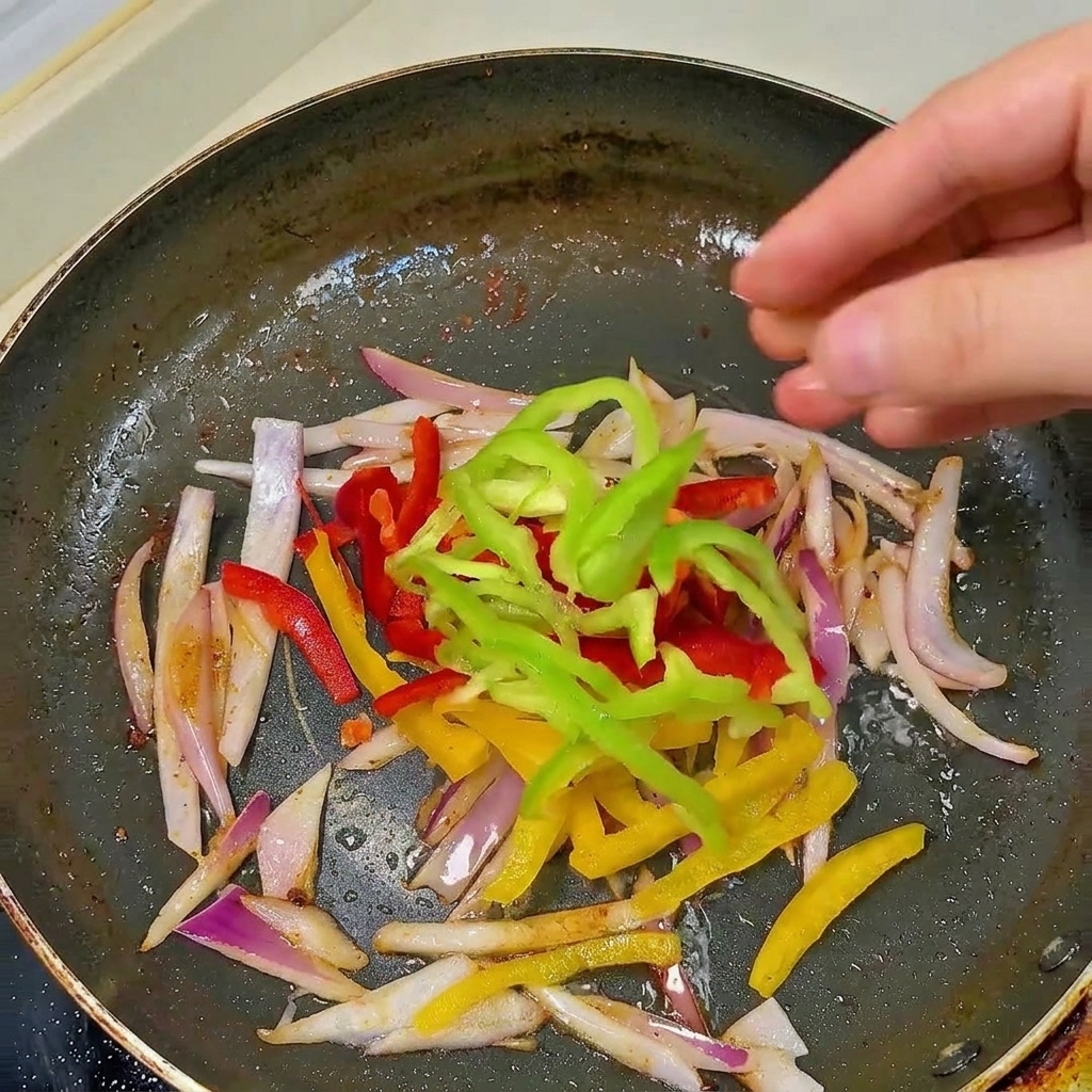 A hand dropping fresh red, yellow, and green bell pepper strips into a black frying pan with sautéed sliced onions.