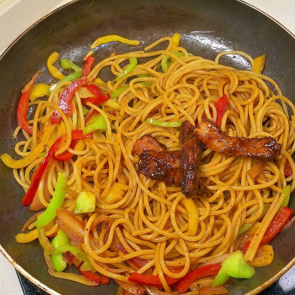 Stir-fried pork strips resting on top of black pepper pasta and bell peppers in a large pan.