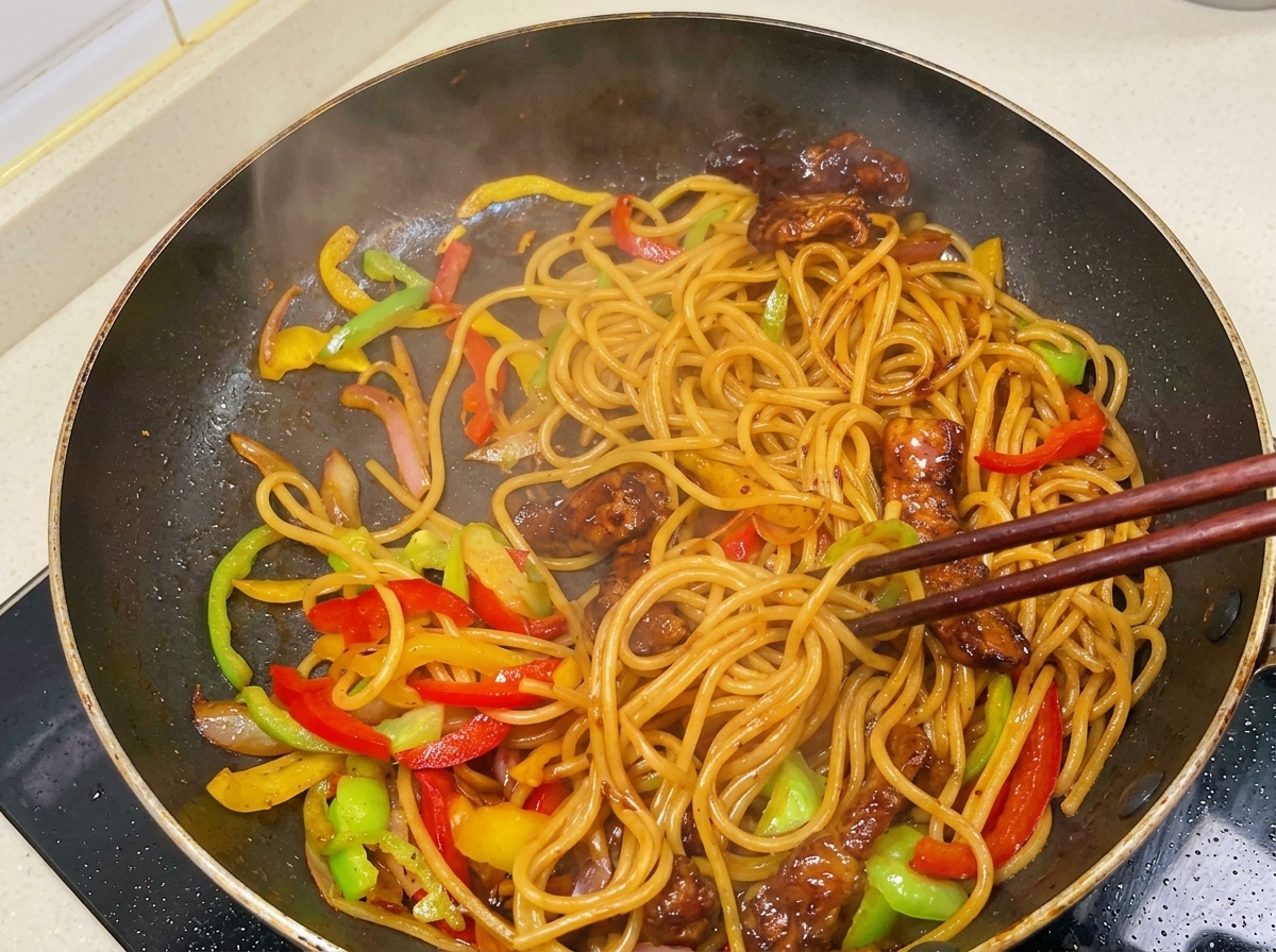 Chopsticks tossing a steaming mixture of spaghetti, seared pork pieces, and sliced bell peppers covered in a dark, glossy sauce.