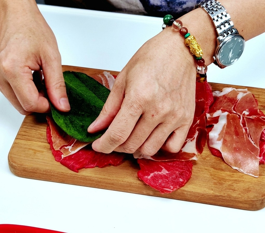 Hands placing a large, fresh green sage leaf on top of a prosciutto-covered veal slice on a wooden board.