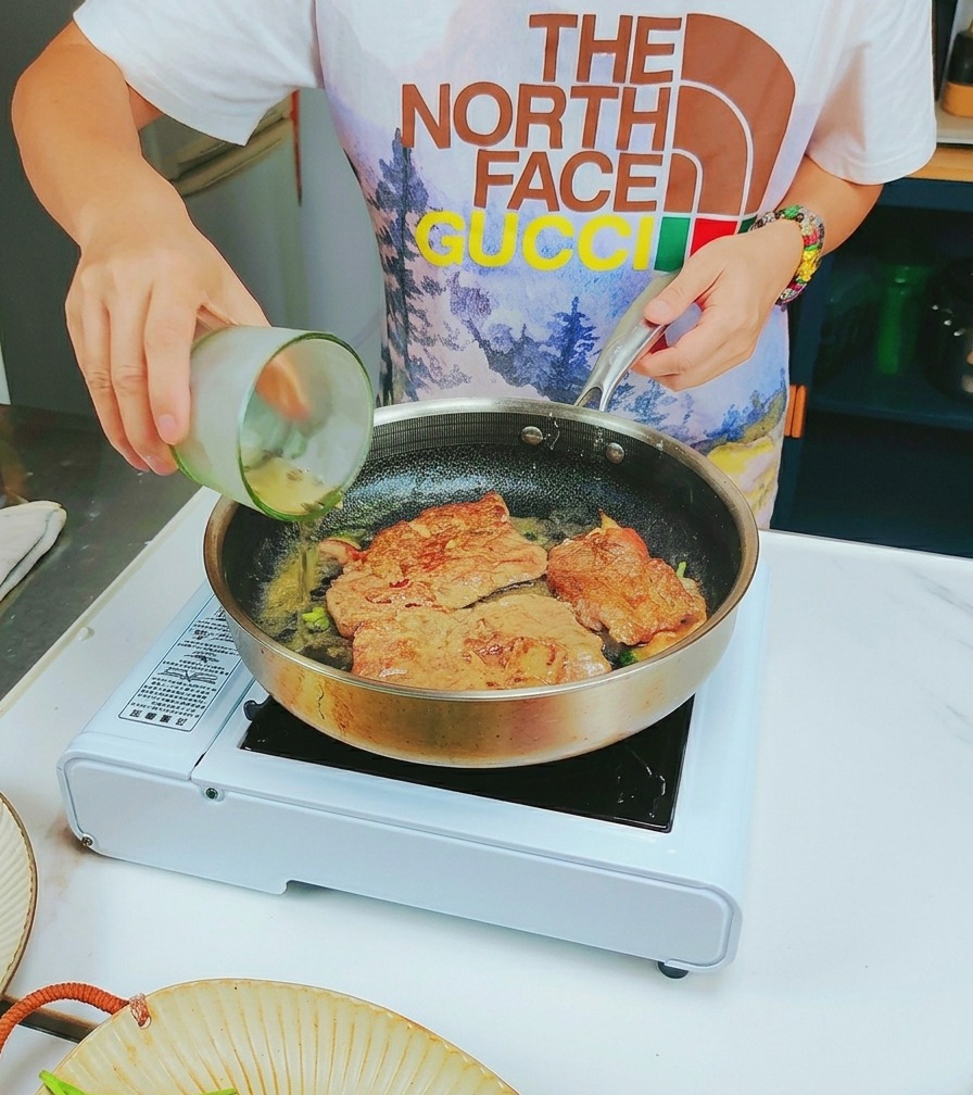 White wine being poured from a glass into a frying pan containing browned veal slices.
