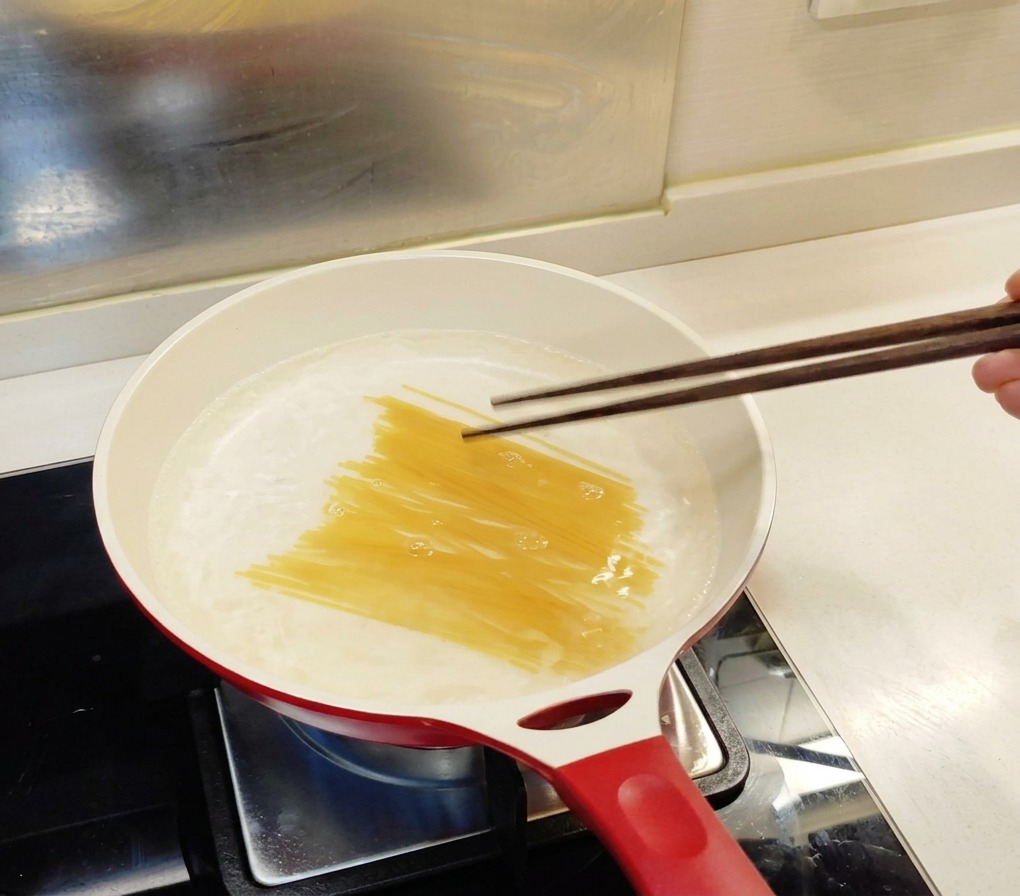 Dry spaghetti being pressed into a white pan of boiling water with dark wooden chopsticks.