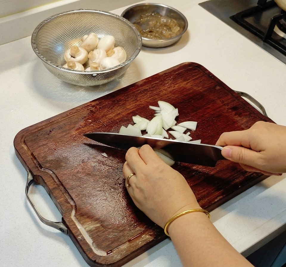 Hands using a chefs knife to finely dice a white onion on a dark wood cutting board next to a bowl of mushrooms.