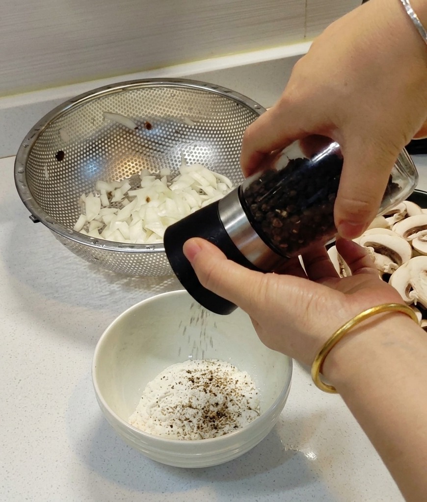 Hands grinding a black pepper mill over a small white bowl filled with white flour.