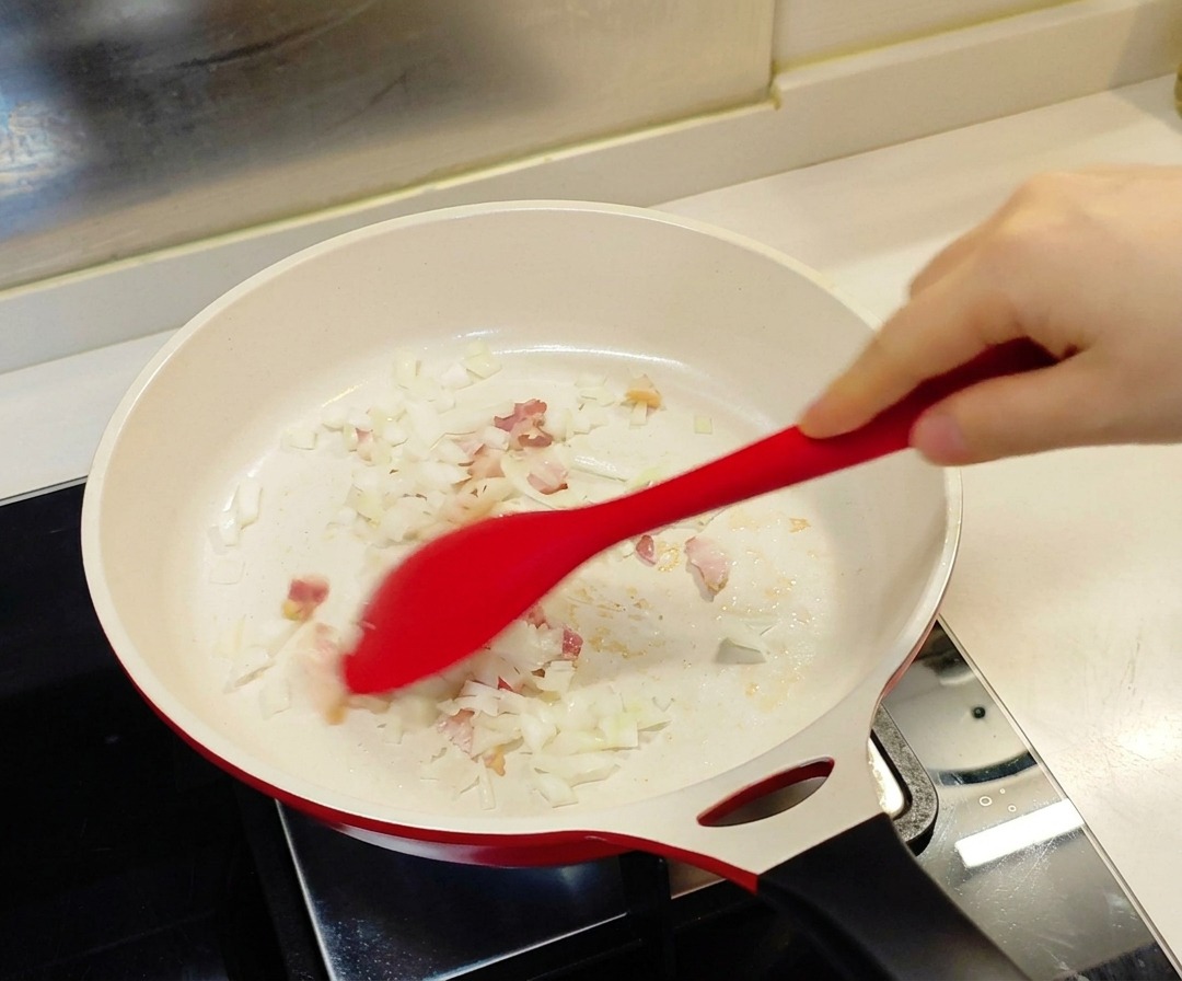 A red spatula stirring chopped onions and small pieces of bacon in a white ceramic-coated pan.