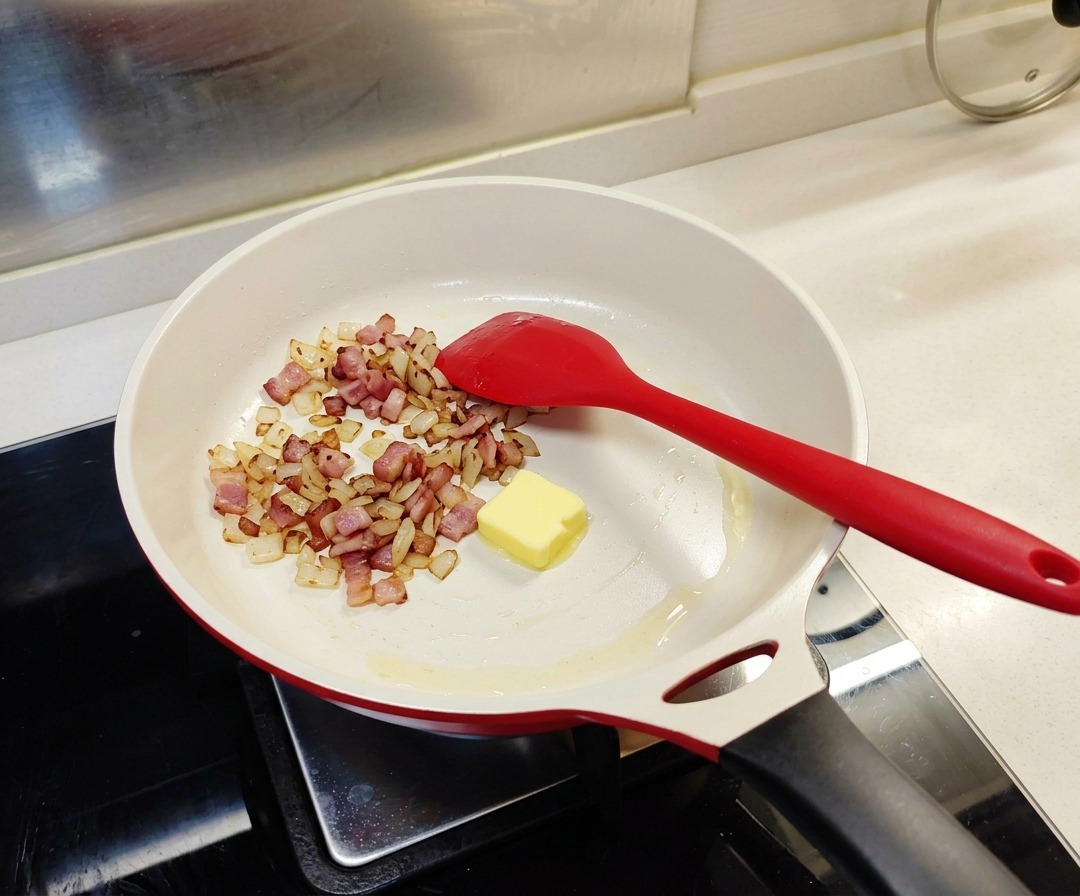 A single square of yellow butter sitting in a white pan with cooked bacon and onions.