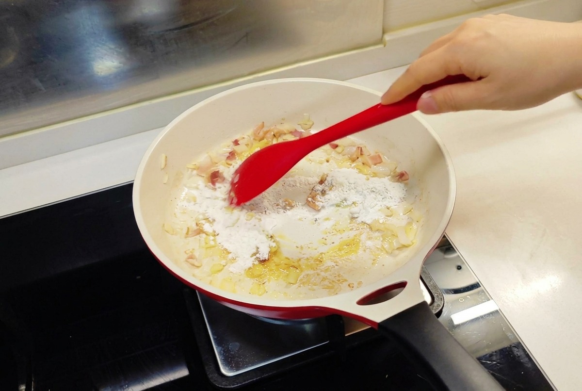A red spatula stirring white flour into a pan containing sautéed onions and melted butter.