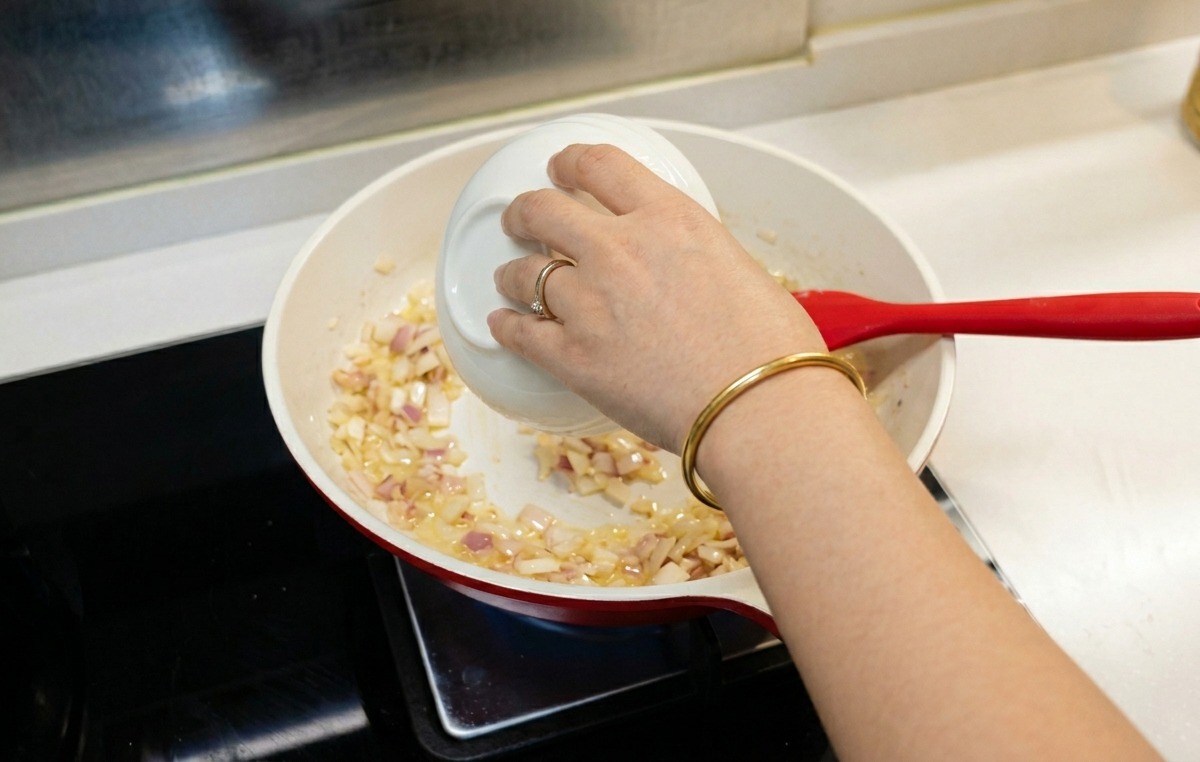 A person pouring water from a small white bowl into a pan with cooked onions and flour.