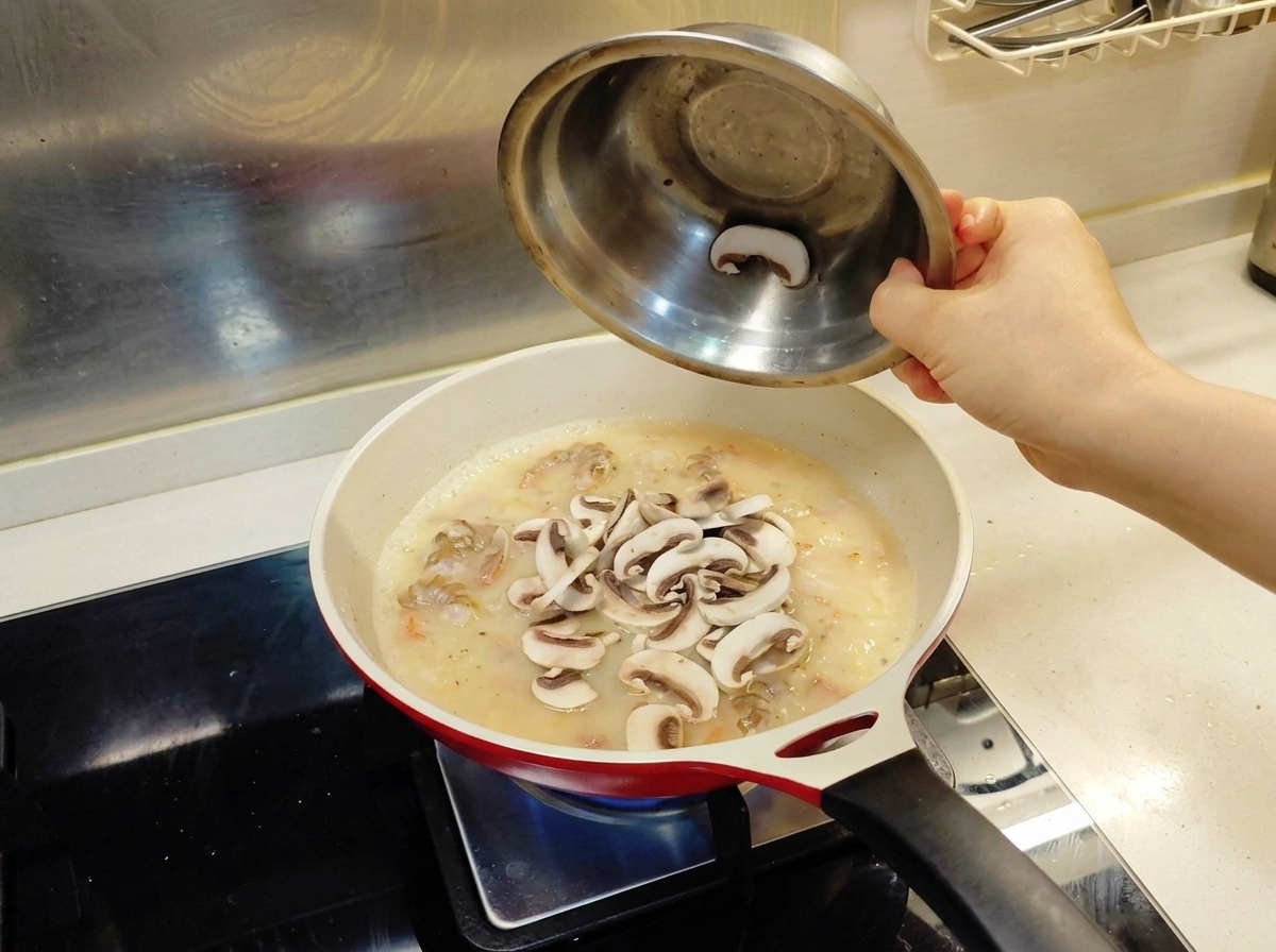 Slices of fresh white mushrooms being poured from a metal bowl into a frying pan containing a creamy sauce base and shrimp.