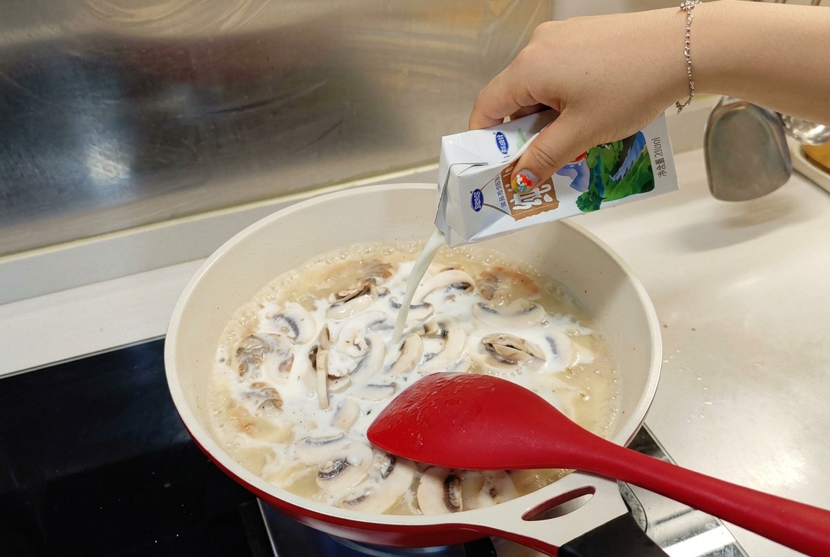 Milk being poured from a white and blue carton into a simmering pan of shrimp and mushrooms.