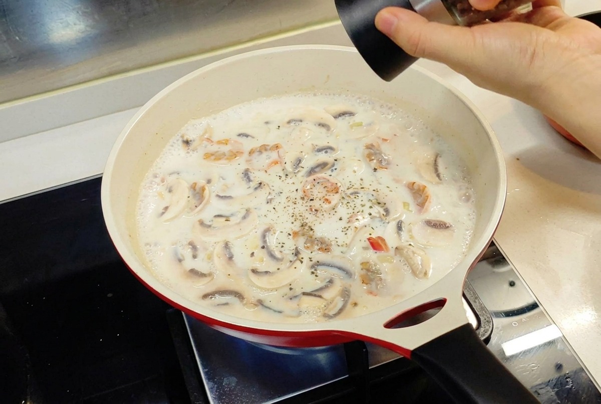 A hand using a pepper grinder to add black pepper to a creamy white sauce simmering in a red pan.