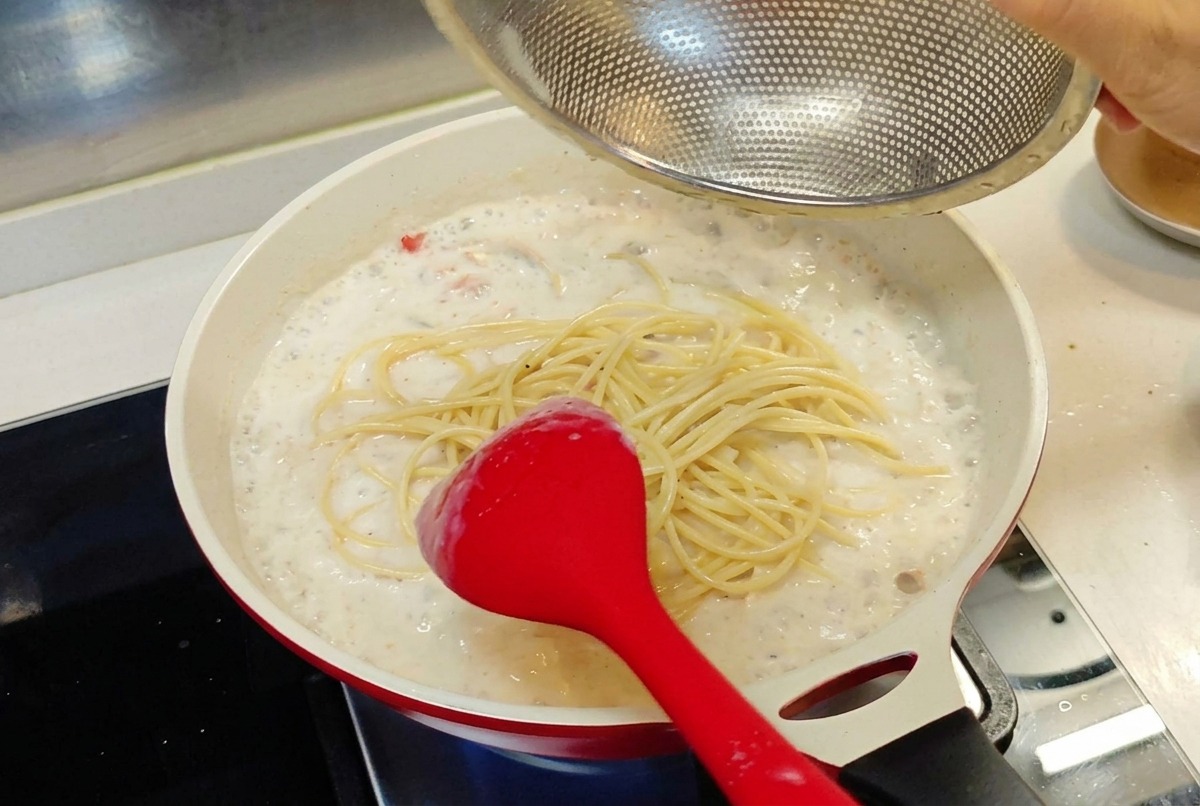 Cooked spaghetti being added from a mesh strainer into a pan of creamy white sauce.
