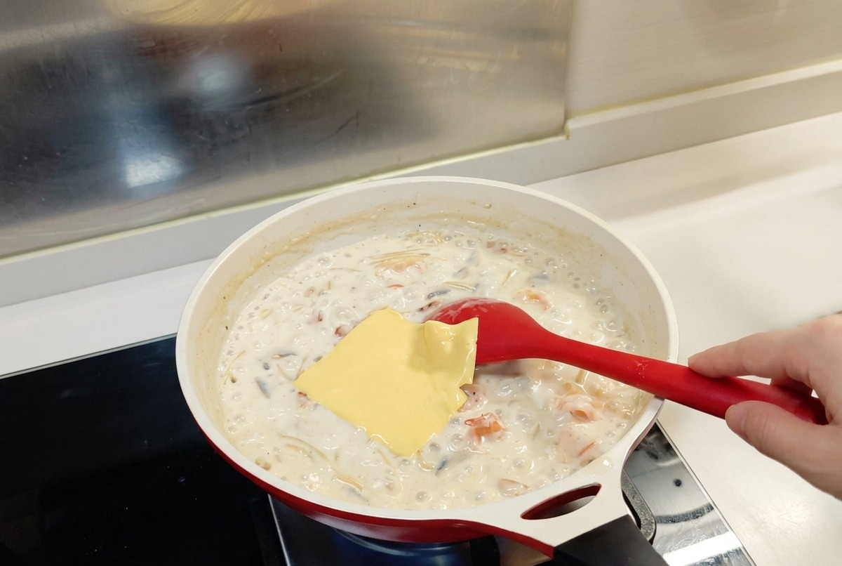 A single square of yellow cheese sitting on top of a bubbling white sauce pasta mixture in a white skillet with a red spoon.