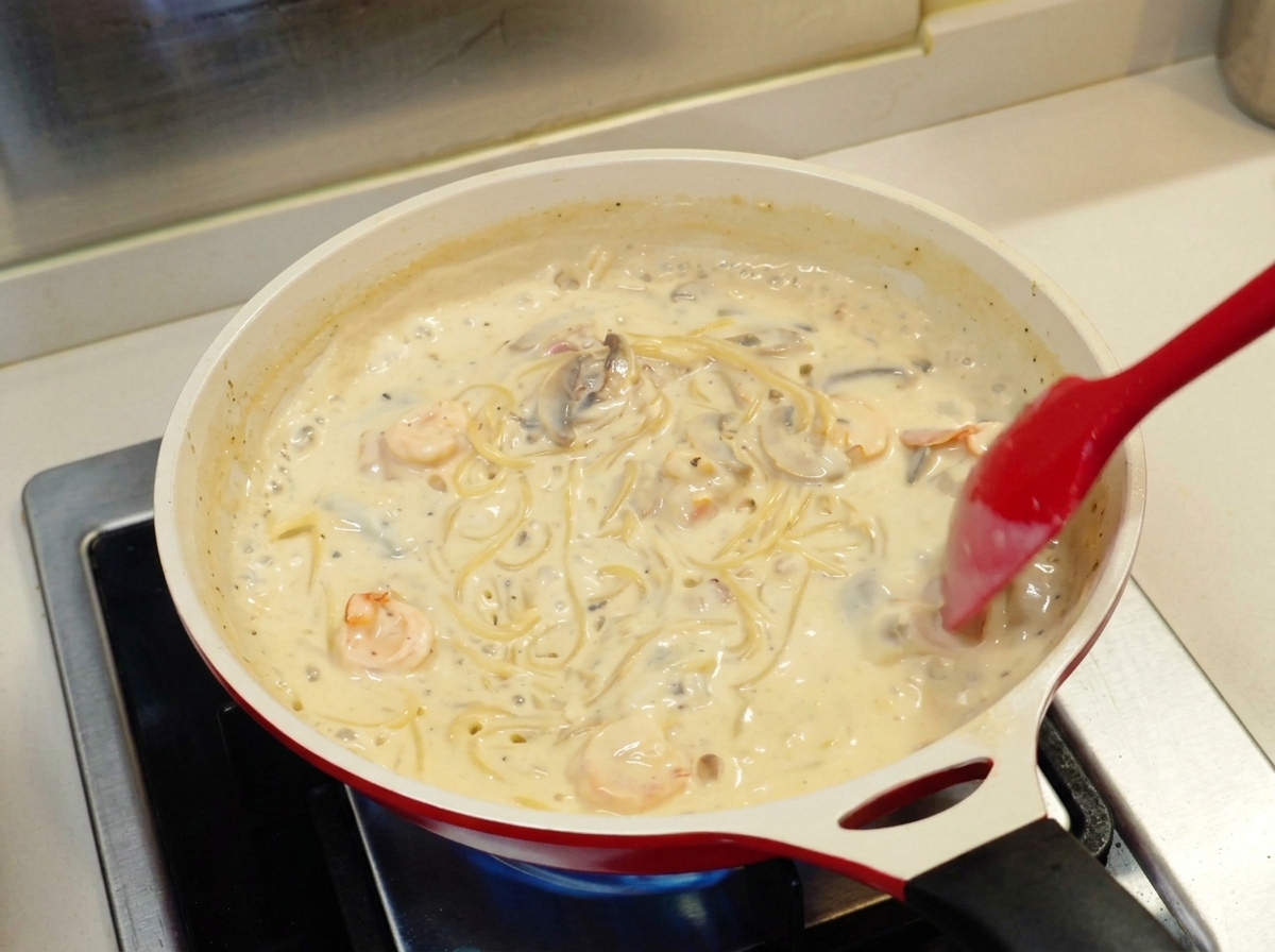 A red spatula stirring spaghetti in a thick, creamy white sauce with shrimp and mushrooms in a white ceramic pan.