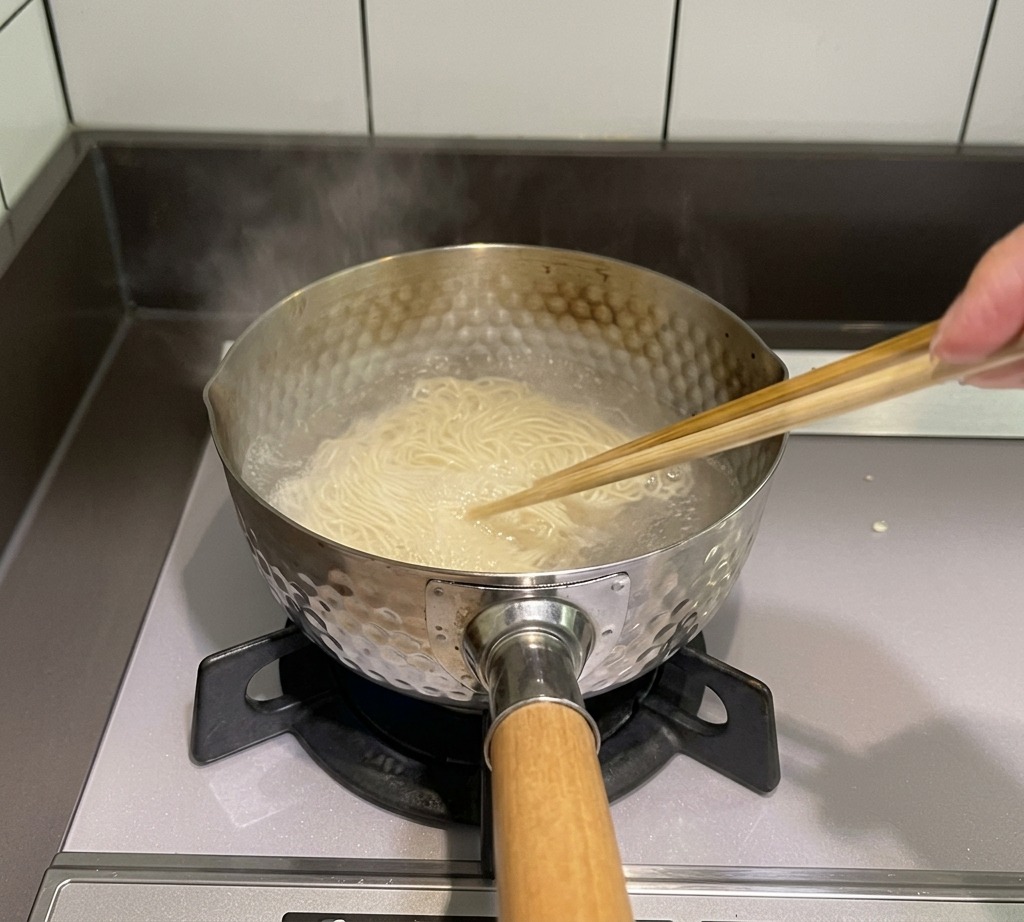 A pair of wooden chopsticks stirring thin noodles in a pot of boiling water on a stove.
