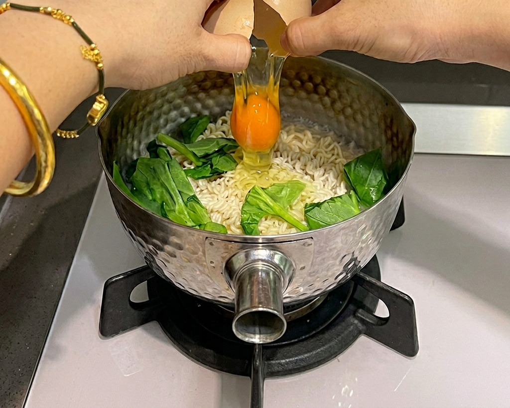 Two hands cracking a raw egg over a pot of boiling instant noodles and green spinach.