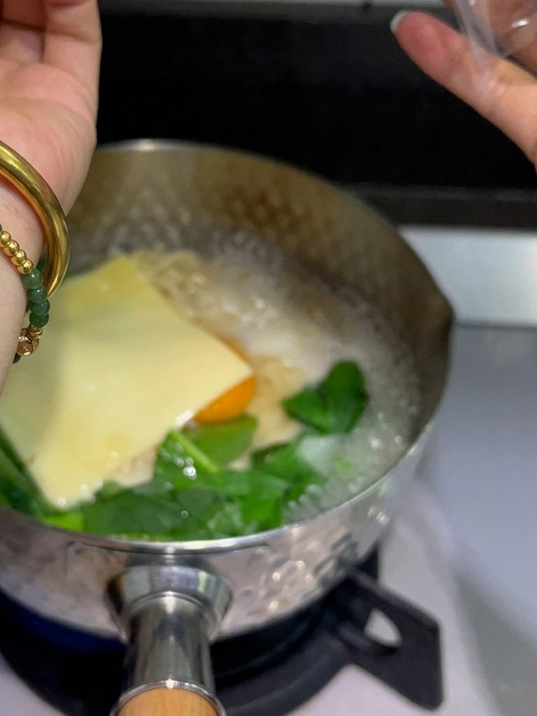 Yellow processed cheese slices being placed on top of an egg and noodles in a steaming pot.