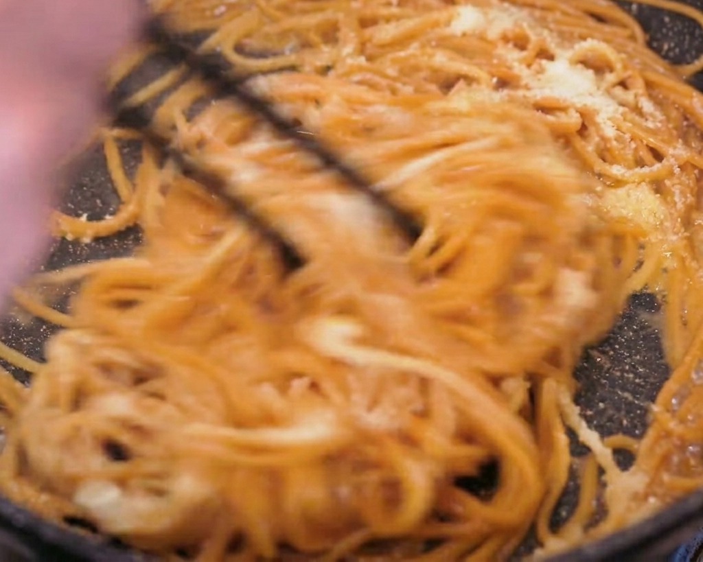 An action shot of whole wheat pasta being tossed with black chopsticks in a pan, showing visible cheese powder and melting butter forming a sauce.