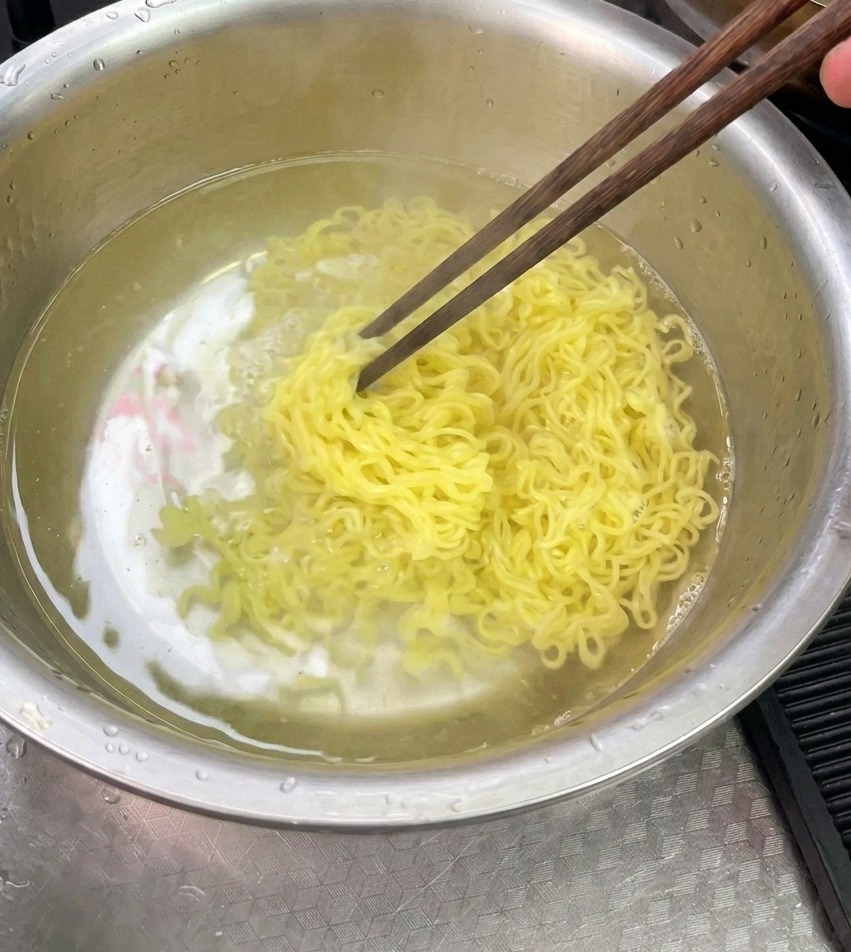 Cooked yellow noodles being rinsed and stirred with chopsticks in a stainless steel bowl filled with cold water.