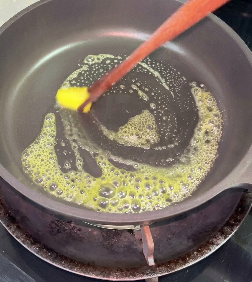 A small square pat of yellow butter melting and bubbling in a black non-stick pan, moved around with wooden chopsticks.