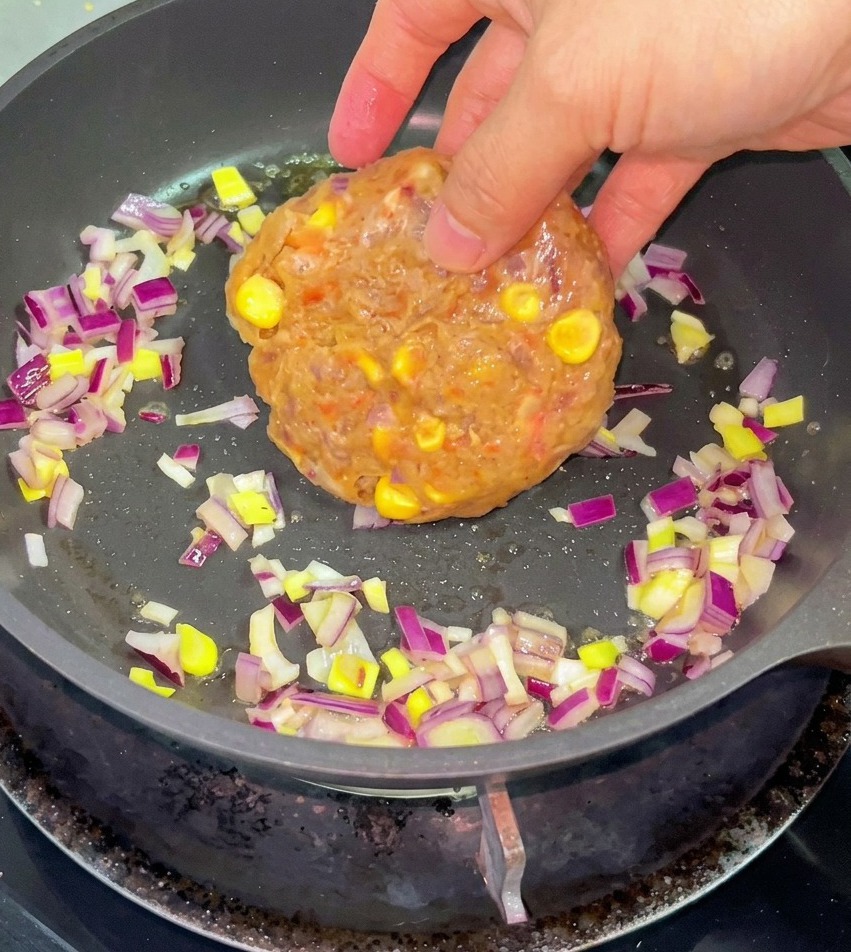 A hand placing a raw beef and corn patty into a frying pan with chopped red onions and melted butter.