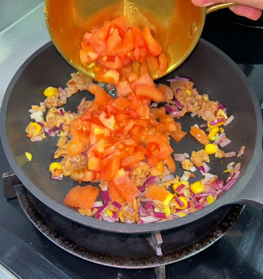 Diced red tomatoes being poured from a small brass bowl into a pan with cooked beef, corn, and red onions.