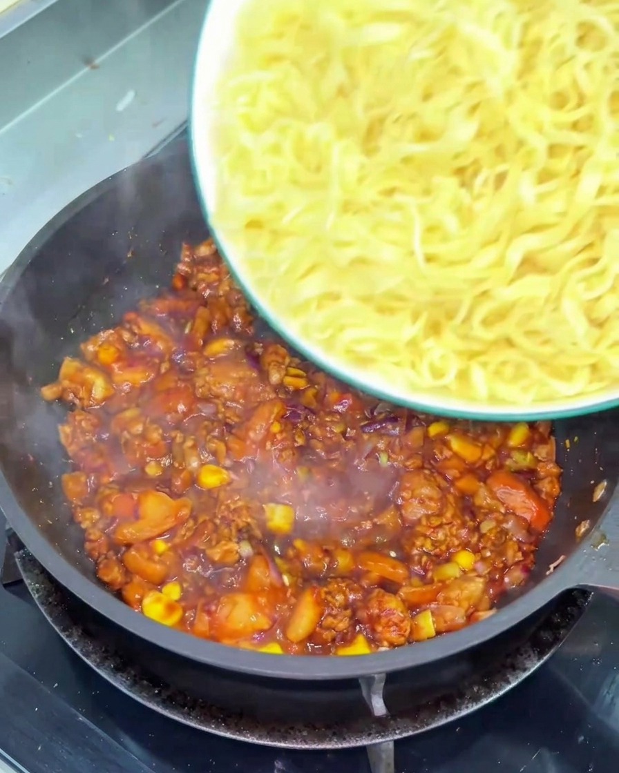 A bowl of wavy yellow noodles being poured into a frying pan filled with a chunky, dark red tomato and beef sauce.