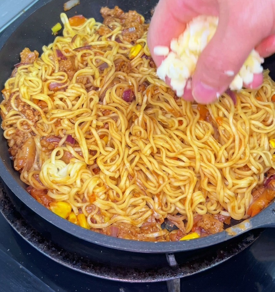 A close-up shot of a hand sprinkling shredded white cheese over a pan of stir-fried noodles mixed with ground beef, onions, and corn.