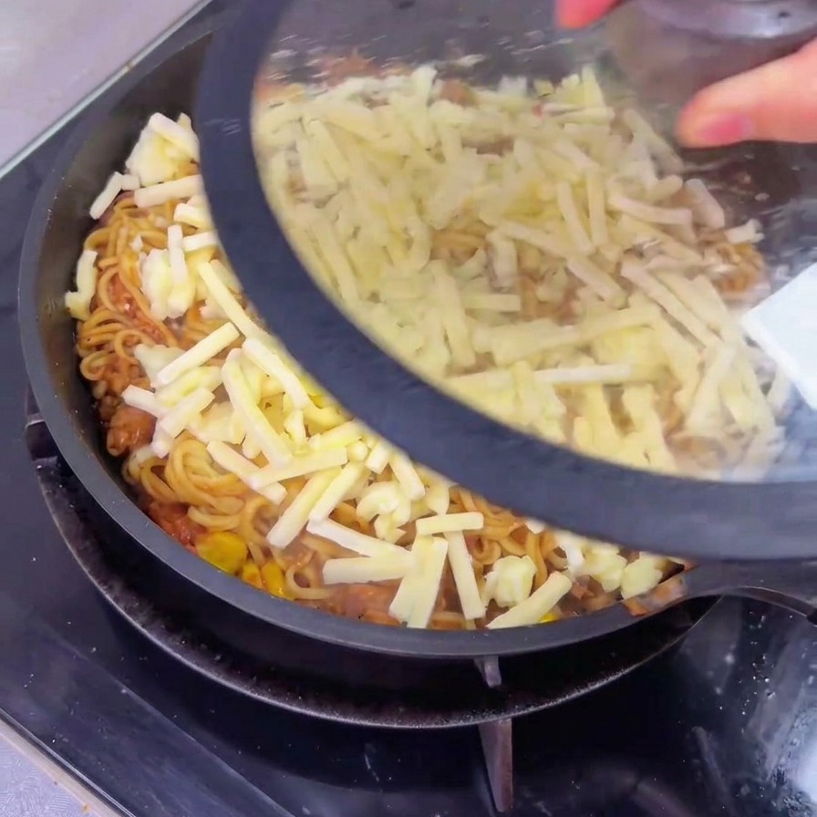 A person placing a clear glass lid onto a large pan containing beef noodles topped with a layer of unmelted shredded cheese.