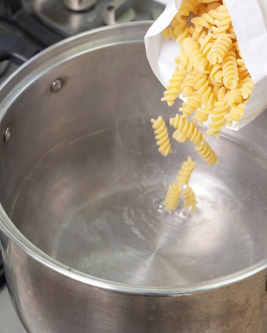 Dry spiral fusilli pasta being poured from a white bag into a large stainless steel pot filled with boiling water.