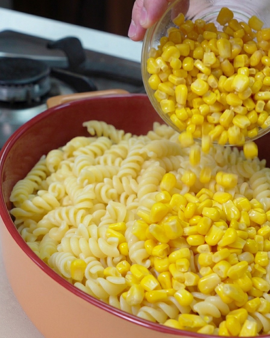 A glass bowl pouring bright yellow sweet corn kernels over a bed of cooked fusilli pasta in a round baking dish.