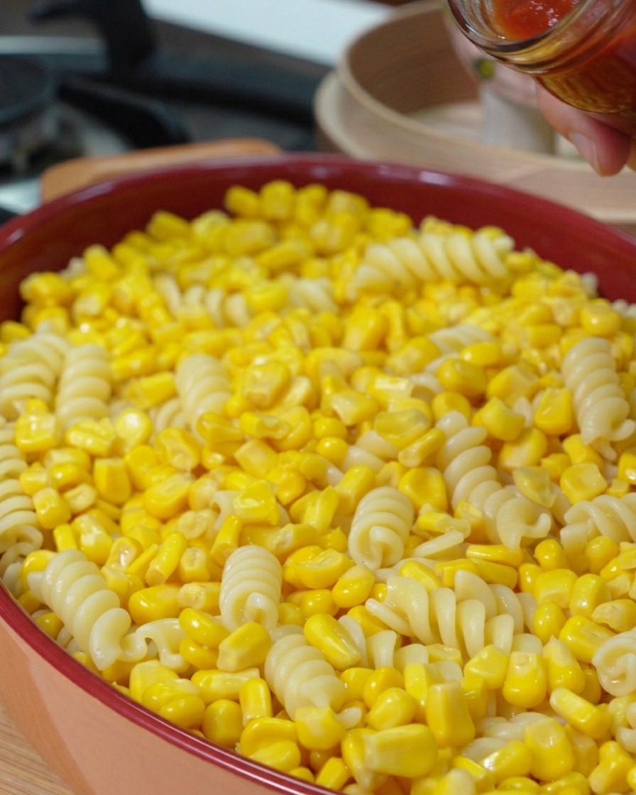 A hand pouring red basil pasta sauce from a glass jar onto a layer of sweet corn kernels and pasta in a baking dish.