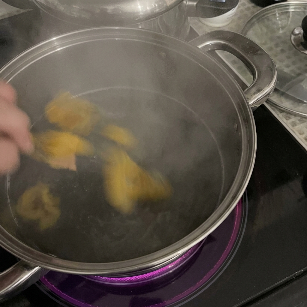A hand adding yellow rigatoni pasta into a stainless steel pot of steaming boiling water on an induction cooktop.