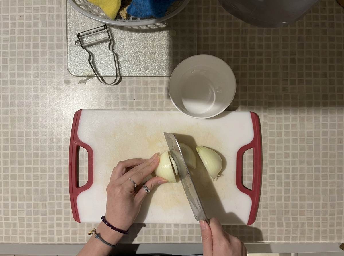 Top-down view of hands using a large knife to dice a white onion on a white cutting board with red handles.