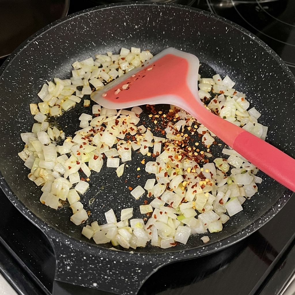 Red dried chili flakes scattered over translucent diced onions in a black frying pan.