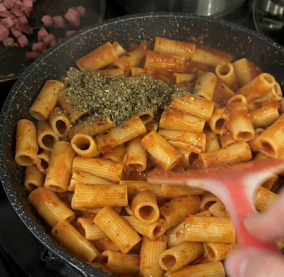 A large pile of dried green basil herb flakes added on top of rigatoni pasta in a tomato sauce inside a black frying pan.