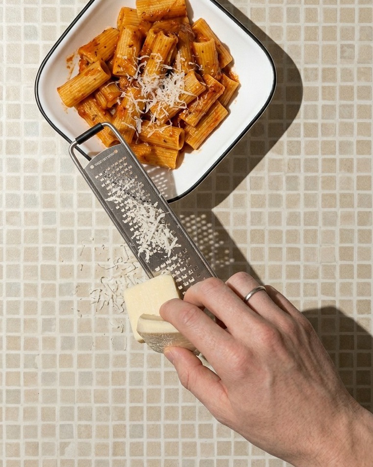 A close-up shot of a hand grating a block of white cheese over a white square bowl filled with rigatoni pasta in a red sauce.