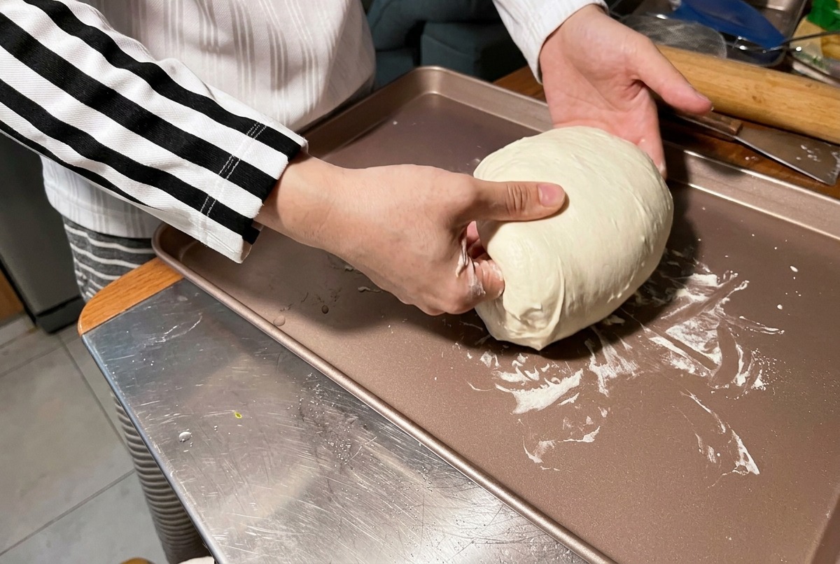A person using their hands to tuck and round a large, soft white dough ball on a bronze-colored baking sheet.