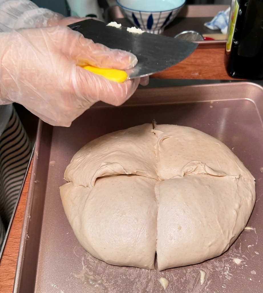 A person wearing a plastic glove using a metal bench scraper to divide a large piece of dough into smaller, rounded sections on a tray.