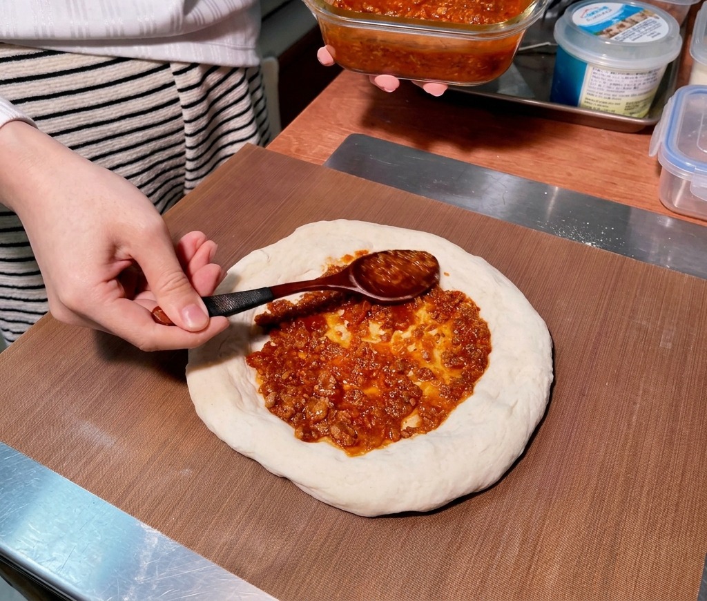 A wooden spoon spreading thick reddish meat sauce in a circular motion on a raw pizza dough base.